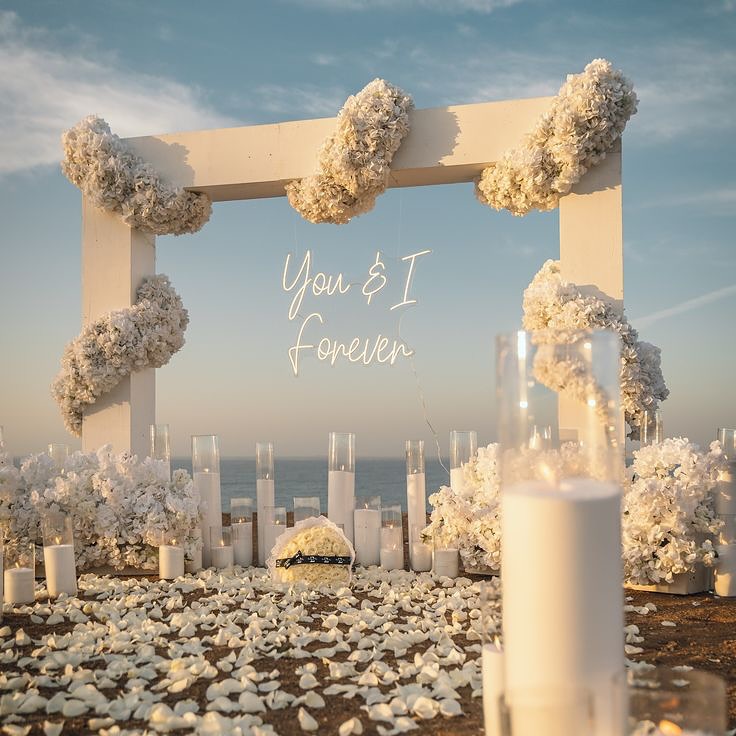 Seaside ceremony arch with white hydrangeas, candles and a 'You & I Forever' neon sign
